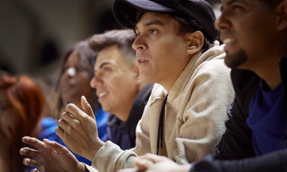 A young man anxiously watches a sporting event surrounded by a crowd of supporters all looking at a field. (Image Credits: Lighthouse Films/Getty Images)