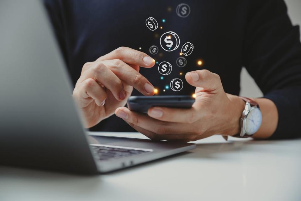 A person with a laptop looks at a mobile device with images of coins appearing on the screen.